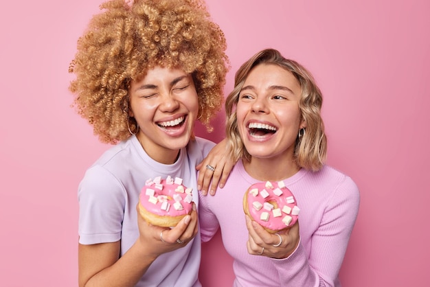 Le donne felici gioiose prendono deliziose ciambelle con marshmallow ridono con gioia non si preoccupano delle calorie si divertono a mangiare il dessert sciocco in giro isolato su sfondo rosa Concetto di cibo malsano