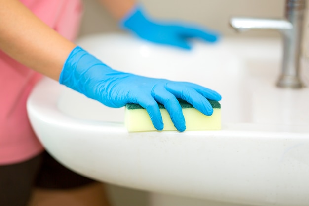 Le donne passano un guanto di gomma blu nella foto, rimuovono e lavano il lavandino del bagno con spray e spugna, concetto di igiene