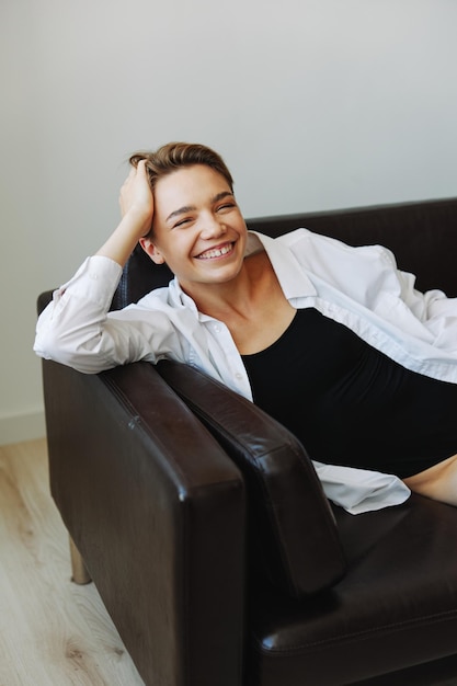 Le donne sdraiate a casa sul ritratto del divano con un taglio di capelli corto in una camicia bianca sorridono la depressione nelle vacanze a casa degli adolescenti