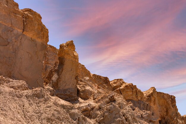 Le formazioni rocciose del paesaggio sono frastagliate e deteriorate, indicando cambiamenti geologici nel tempo. Il cielo è adornato da nuvole morbide illuminate in tonalità di rosa e blu, creando un'atmosfera eterea.