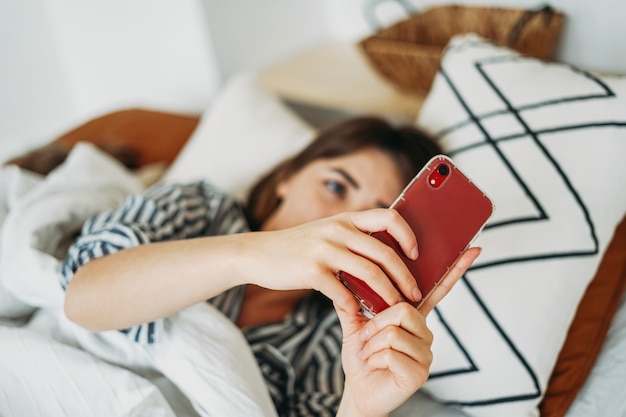 Le free lance giovani lavoranti della donna castana in abbigliamento casual facendo uso del cellulare svegliano a letto a casa nel soggiorno interno luminoso a casa