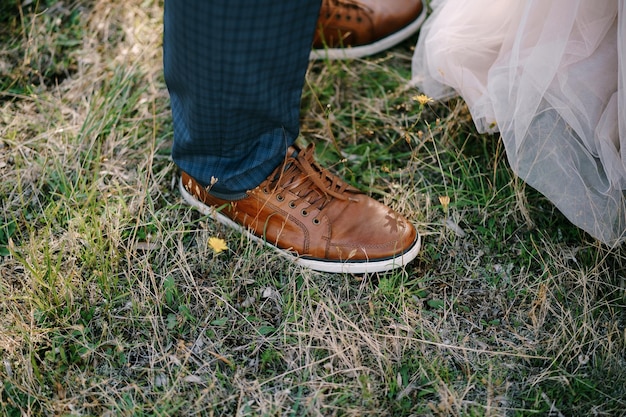 Le gambe dello sposo in scarpe da ginnastica marroni stanno sull'erba verde vicino al primo piano del vestito dalla sposa
