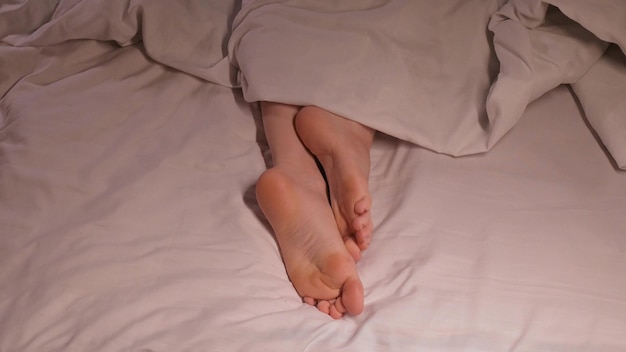 le gambe e le dita delle gambe femminili prurito e scabbia di notte a letto su un lenzuolo sotto una coperta donna ragazza a piedi nudi piede con funghi sotto stress dorme a letto in una camera da letto di notte a casa