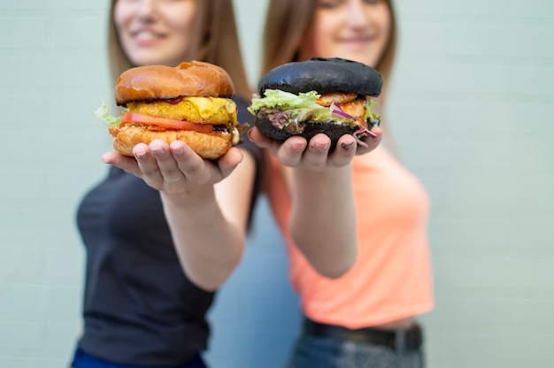 Le giovani belle studentesse stanno vicino al muro blu, tengono gli hamburger e sorridono, sono felici e mostrano i cheeseburger sulla macchina fotografica