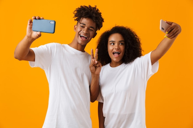 Le giovani coppie africane sveglie felici isolate sopra lo spazio giallo che mostra il gesto di pace fanno selfie dai telefoni cellulari.