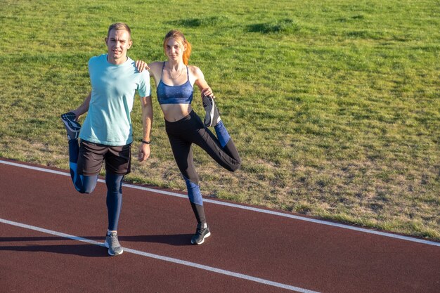 Le giovani coppie degli sportivi adatti ragazzo e ragazza che fanno scaldarsi si esercitano prima di correre sulle piste rosse dello stadio pubblico all'aperto.