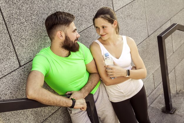 Le giovani coppie sportive stanno riposando e controllano insieme il loro orologio fitness, bevendo acqua. concetti sportivi e tecnologici.