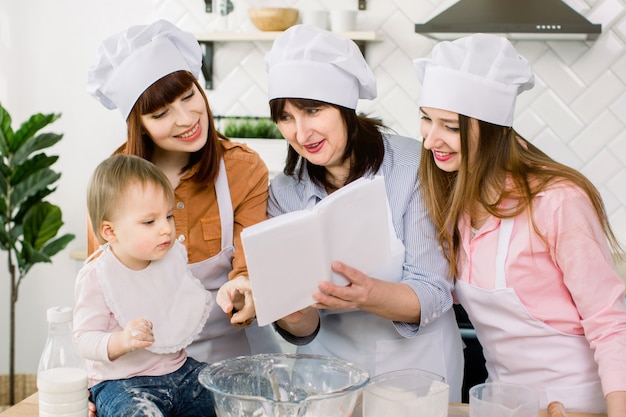 Le giovani donne attraenti, la donna di mezza età e la piccola figlia carina stanno cucinando sulla cucina. Divertirsi insieme mentre si fanno torte e biscotti. Le donne leggono il libro di cucina con ricette