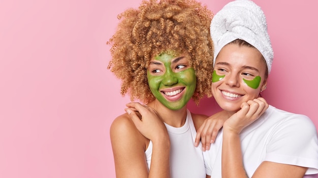 Le giovani donne positive applicano la maschera di bellezza e i cerotti in idrogel sotto gli occhi per il trattamento della pelle coccolano il complesso dopo aver fatto la doccia a stretto contatto l'uno con l'altro sorridono felicemente isolati sul muro rosa