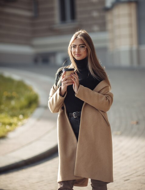 Le giovani donne vestite con un cappotto alla moda bevono caffè su uno sfondo di architettura urbana