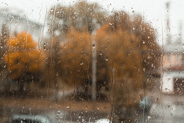 Le gocce di pioggia scendono sulla superficie della finestra, la città e gli alberi gialli sono sfocati, copia dello spazio. Giornata nuvolosa e piovosa autunnale. Concetto di maltempo.