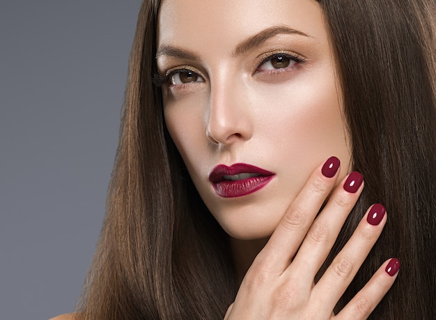 Le labbra rosse della bella donna castana manicure con il primo piano naturale del fronte di trucco dei capelli lunghi. Colpo dello studio. Colore di sfondo.