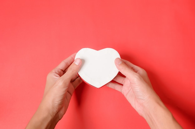 Le mani che tengono un cuore bianco modellano il contenitore di regalo su un fondo rosso. Confezione regalo di San Valentino.