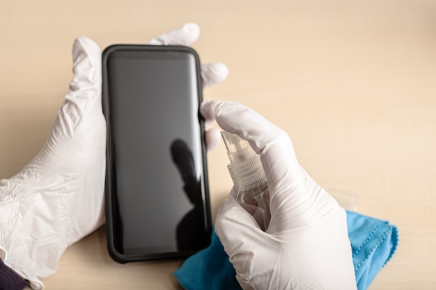 Le mani con i guanti puliscono il telefono cellulare con spray disinfettante. COVID-19 Concetto di prevenzione della contaminazione da epidemia di coronavirus.