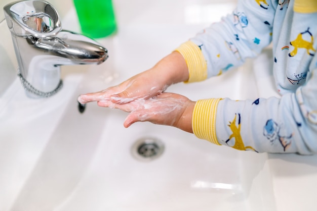 Le mani dei bambini in pigiama si lavano le mani con molta schiuma in un lavandino del bagno con un rubinetto