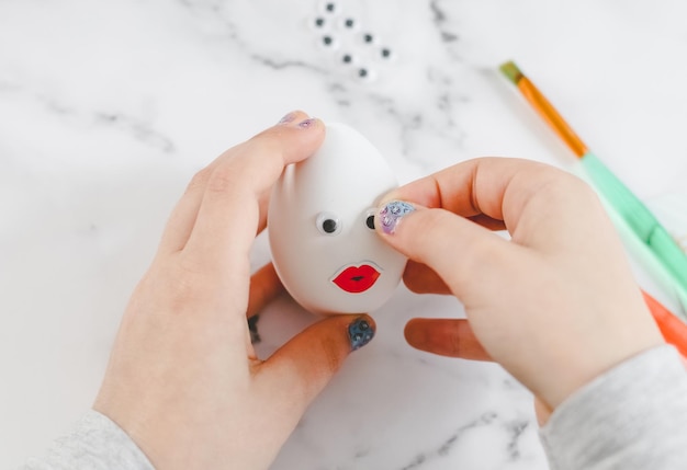 Le mani dei bambini tengono un uovo bianco e incollano gli occhi decorativi adesivi seduti a un tavolo di marmo