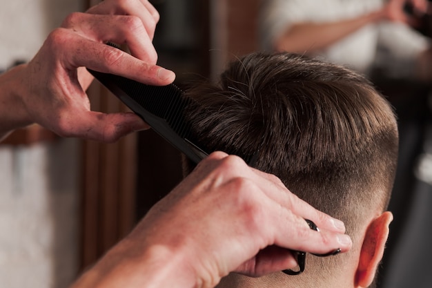 Le mani del barbiere che fanno taglio di capelli all'uomo attraente nel negozio di barbiere