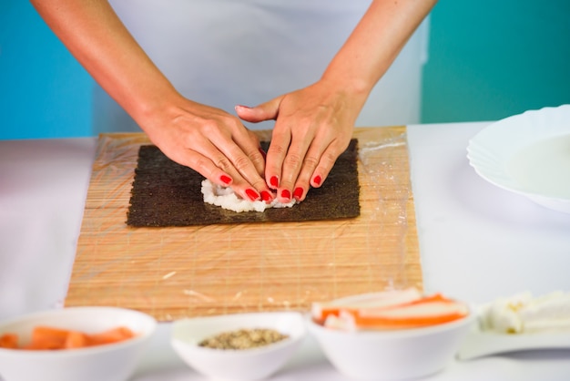 Le mani del cuoco unico della donna che riempiono i rotoli di sushi giapponesi con riso su uno strato dell&#39;alga di nori.
