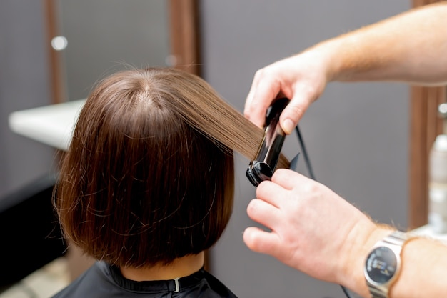 Le mani del parrucchiere stanno raddrizzando i capelli della donna con lo strumento raddrizzatore nel parrucchiere, vista posteriore.