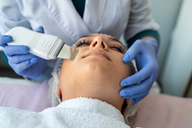 Le mani dell'estetista che utilizzano il dispositivo per la pulizia ad ultrasuoni facendo la procedura della donna nella clinica di cosmetologia