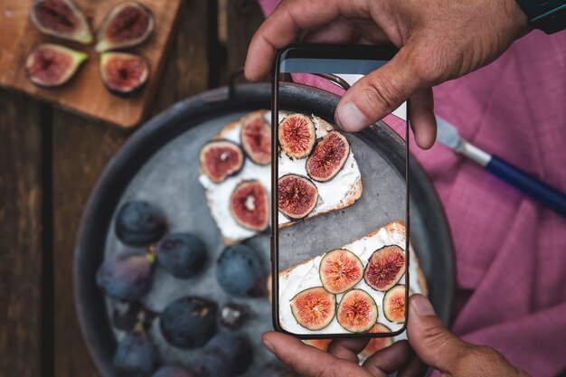 Le mani dell'uomo scattano una foto del cibo sul tavolo con il telefono. Colazione. Panino con ricotta morbida e fichi. Foto di smartphone per social media o post sul blog. Vegetariano, sano, biologico