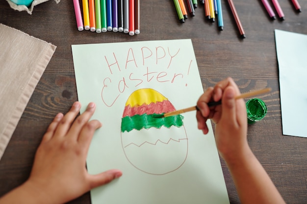 Le mani della bambina di razza mista che dipingono un grande uovo di Pasqua su carta bianca tra evidenziatori colorati e pastelli mentre sono seduti da un tavolo di legno