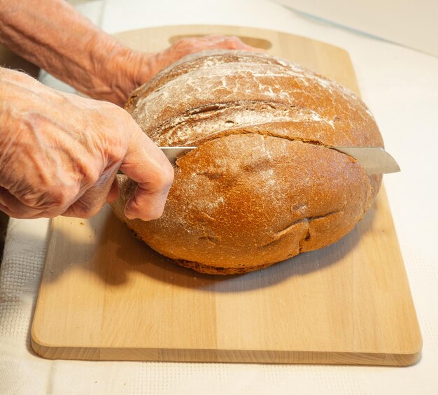 Le mani della donna anziana tagliano il pane di segale rotondo nella farina sul bordo della cucina