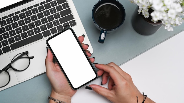 Le mani della donna che tengono il telefono cellulare con lo schermo bianco alla sua scrivania dell'ufficio