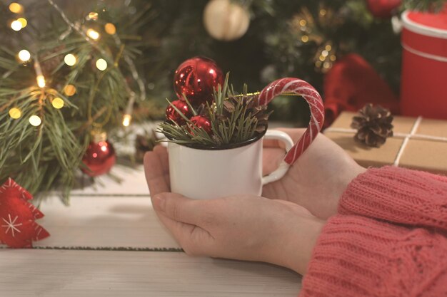 Le mani della donna che tengono tazza con palloncini rossi rami di albero di Natale e lecca-lecca sul tavolo bianco