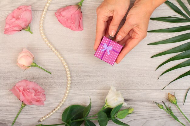 Le mani della donna che tengono una confezione regalo su fondo di legno grigio con bellissimi fiori e foglie verdi. Concetto di fare un regalo nei giorni festivi. Vista dall'alto.