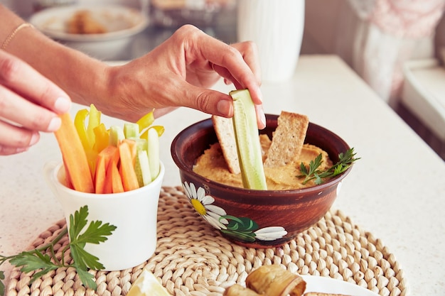 Le mani della donna immergono il cetriolo fresco in hummus fatto in casa Biscotti prezzemolo e verdure fresche sul tavolo Spuntini rustici vegetariani sani da vicino