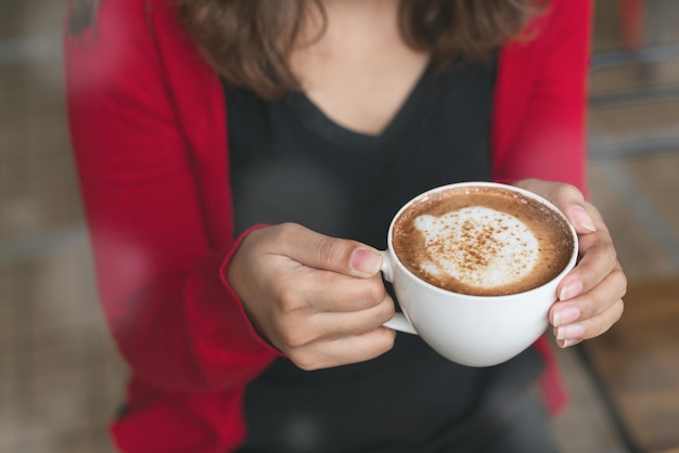 Le mani della donna in cadigan rosso che tiene tazza del caffè di cappucino nell&#39;inverno.