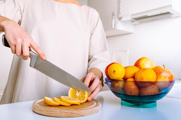 le mani della donna tagliano le fette di limone sul tagliere di legno per preparare una bevanda rinfrescante al limone, una ciotola di fresco