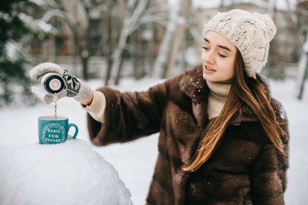 Le mani della donna versano il tè o il caffè caldo dal thermos sulla ragazza del fondo della foresta di inverno che usa un thermos su una tazza rossa della montagna innevata