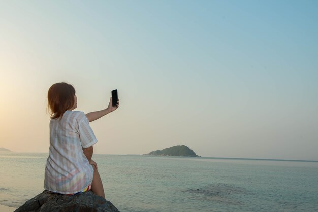 Le mani della giovane donna usano lo smartphone seduto su una roccia godendosi la spiaggia