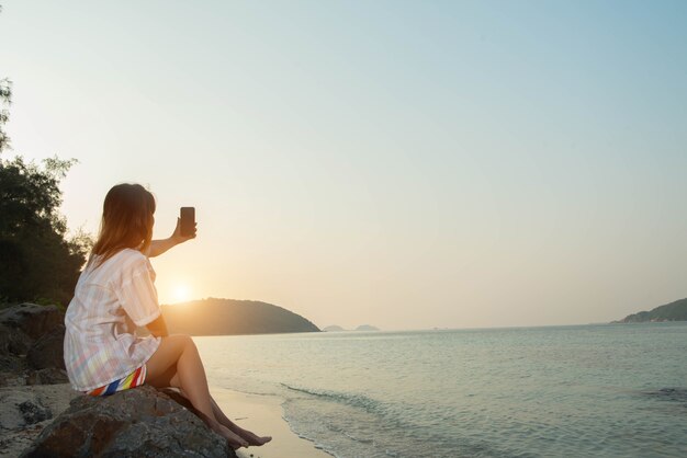 Le mani della giovane donna usano lo smartphone seduto su una roccia godendosi la spiaggia