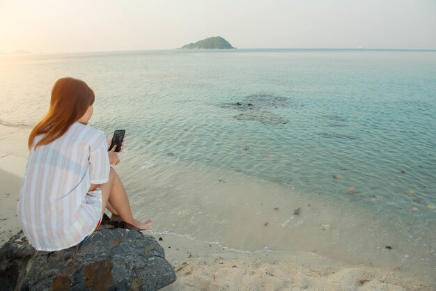 Le mani della giovane donna usano lo smartphone seduto su una roccia godendosi la spiaggia