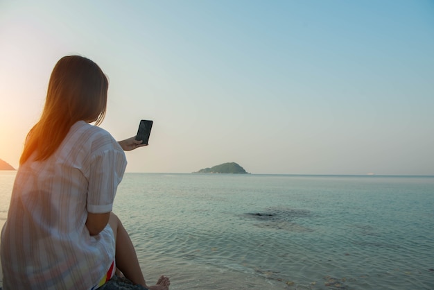 Le mani della giovane donna usano lo smartphone seduto su una roccia godendosi la spiaggia