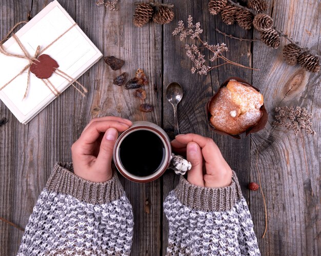 Le mani delle donne in un maglione marrone a maglia che tiene una tazza di ceramica con caffè nero