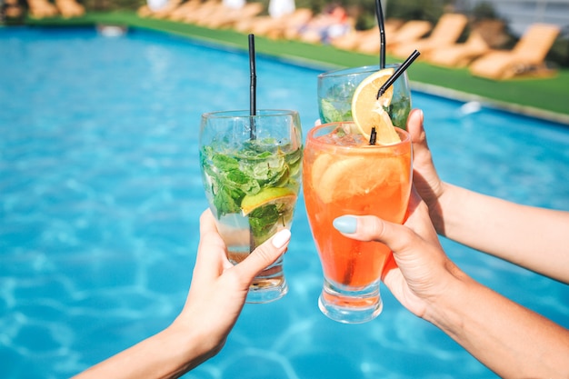 le mani delle donne tengono tre bicchieri di cocktail davanti alla piscina