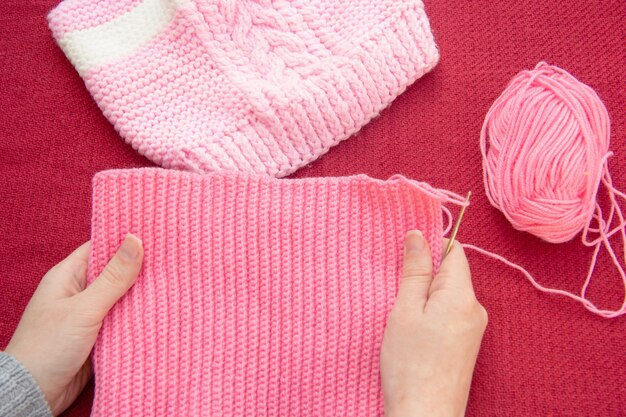 Le mani delle donne tengono un panno rosa lavorato a maglia, che è legato con un uncinetto. Nelle vicinanze c'è un cappello da bambino legato con dei raggi.