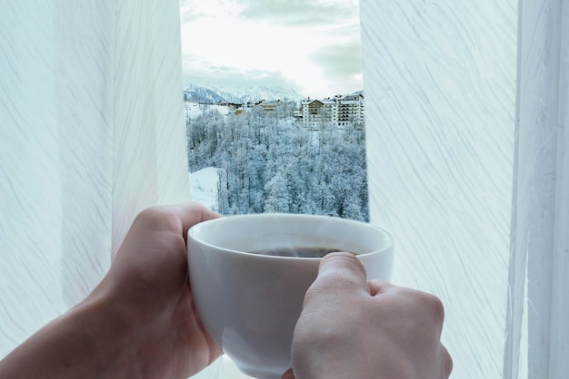 le mani delle persone tengono una tazza di caffè caldo di fronte alla finestra nella stagione invernale b