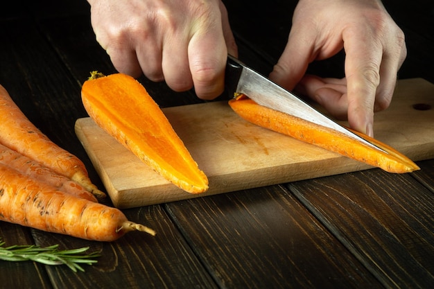 Le mani dello chef con un coltello tagliano carote crude fresche su una tavola da taglio di cucina prima di preparare un piatto vegetariano con verdure
