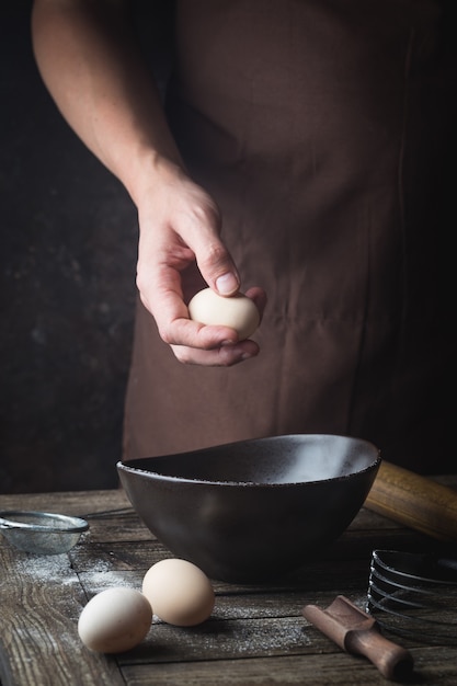 Le mani dello chef professionista stanno rompendo un uovo nella ciotola per fare l'impasto su un tavolo di legno, al buio
