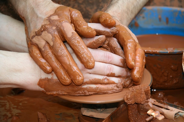 Le mani di un'artigiana che guidano un'altra mano, per insegnargli a lavorare con la ruota in ceramica.