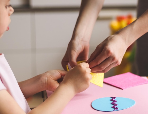 Le mani di una bambina caucasica tengono un adesivo giallo e le mani della mamma aiutano a staccare una striscia per le uova di feltro