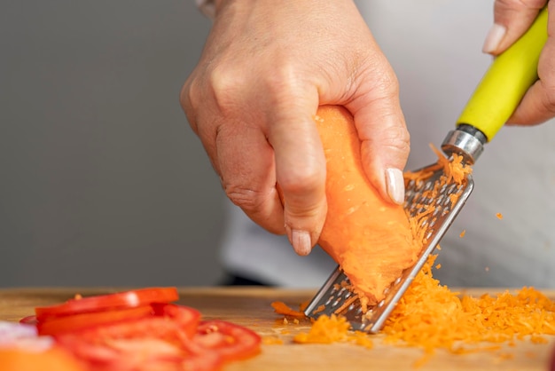 Le mani di una donna adulta che grattugiano una carota con una grattugia su una tavola di legno per la preparazione delle lasagne