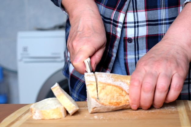 Le mani di una donna anziana tagliano baguette di pane bianco con un coltello in cucina su un tavolo marrone senza un primo piano del viso