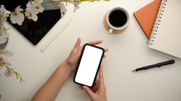 Le mani di una ragazza che tengono il mockup dello schermo vuoto dello smartphone su uno spazio di lavoro minimo. vista dall'alto