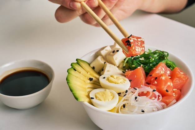 Le mani femminili che tengono le bacchette colpiscono la ciotola con salmone, salsa di soia del sesamo dell&#39;avocado